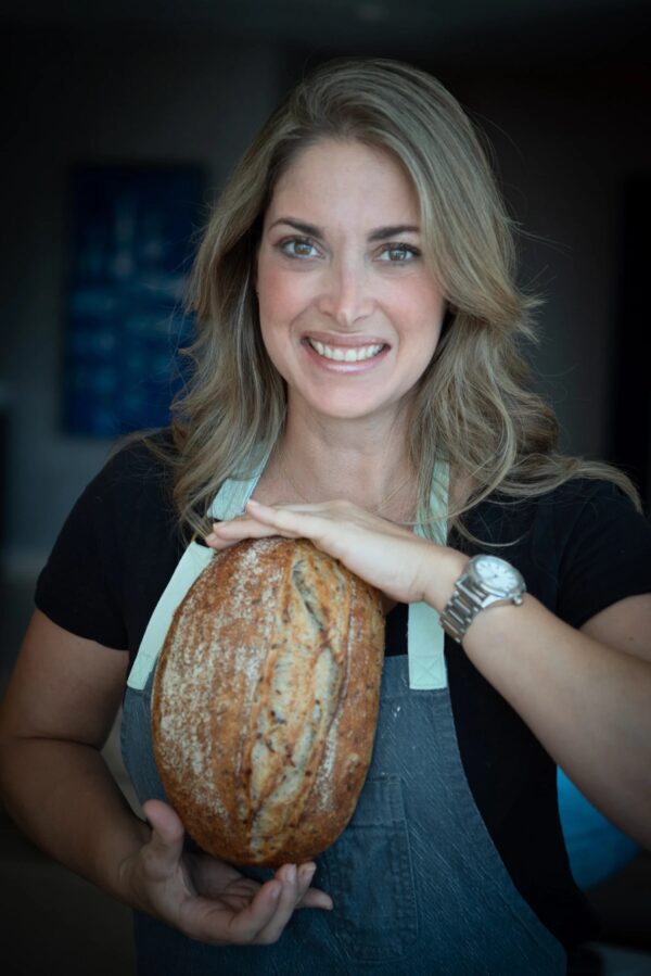 Chef Edna Cochez holding a loaf of fresh baked bread, wearing a black short sleeve top and her chef's apron, smiling