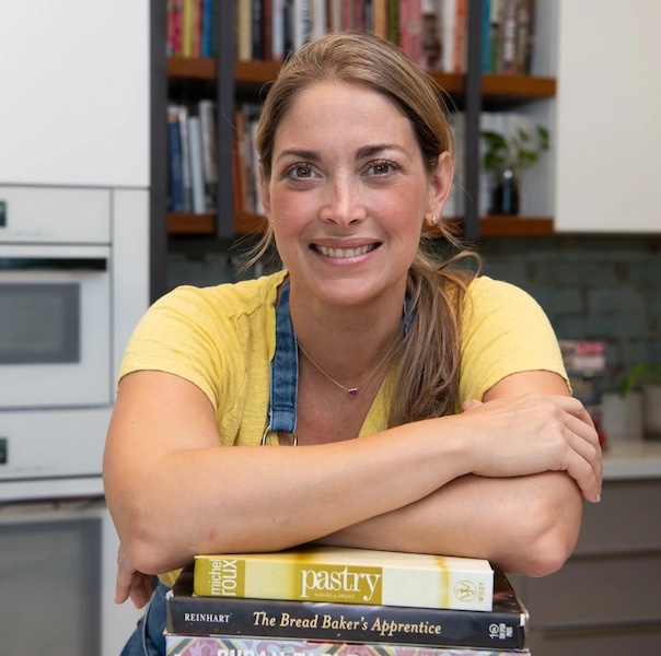 Chef Edna Cochez with arms resting on a stack of recipe books, wearing a yellow short sleeve shirt and her chef's apron, smiling with hair in a ponytail.