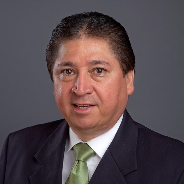 Mike Aguilera in dark blazer, white shirt and green tie on gray background.